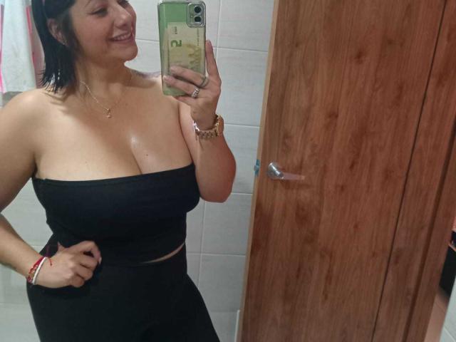 AngeleRoux trägt ein schwarzes, trägerloses Top, das ihre großen Brüste zeigt, und schwarze Hosen. Sie macht ein Selfie vor einem Spiegel mit einem grünen Handy und lächelt. Im Hintergrund ist eine Holztür zu sehen..