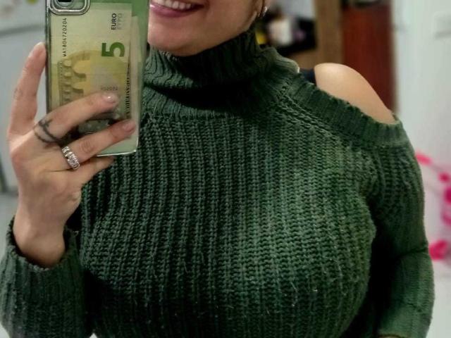 AngeleRoux, con senos grandes, vistiendo un suéter verde de cuello alto de punto, sosteniendo un teléfono, sonriendo, mostrando un anillo y llevando un anillo en su dedo..