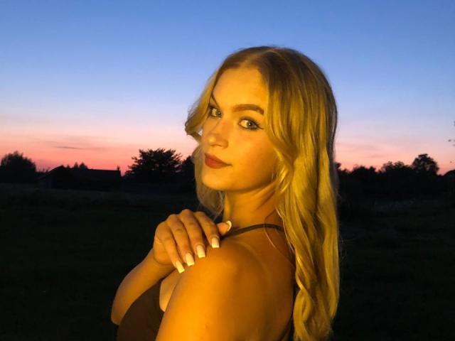 AlinaLemon with long blonde hair, looking over her shoulder, has her hand on her shoulder, showing her tits, against a sunset sky. Her nails are painted white.