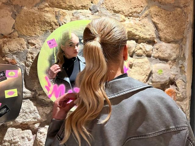 AlinaLemon with long blonde ponytail, wearing grey denim jacket, looking at neon green mirror with pink sticky notes, holding pink sticky note, in stone-walled room.