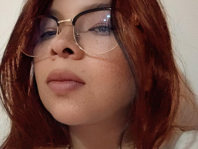 Unicornia69 with red hair, wearing glasses, has freckles and slightly parted pink lips, looking up with a subtle pout, in a selfie. Her face is the focus.