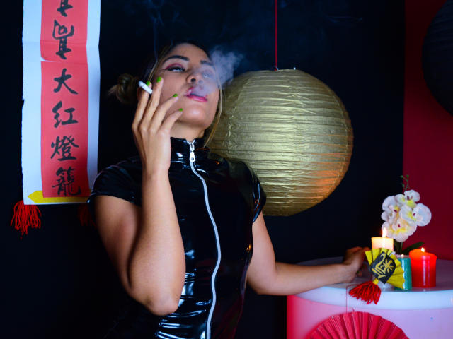 SteffyMiller, avec des ongles verts, fumant une cigarette, expirant de la fumée, portant une robe en latex noire, tenant son menton, un lanterneau en papier doré en arrière-plan, des bougies rouges, des orchidées blanches et un éventail rouge..