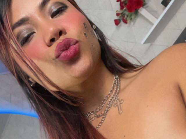 SteffyMiller pouts with glossy pink lips, brown eyes, and a chain necklace with a cross, red-brown hair, and small facial marks. Her bare shoulders are visible.