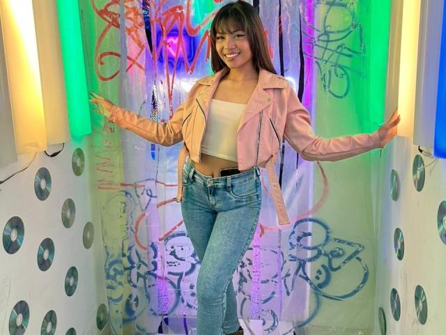 SteffyMiller with medium tits, smiling, wearing a pink leather jacket, white crop top, and blue jeans, standing with arms out, in a colorful, graffiti-covered space.