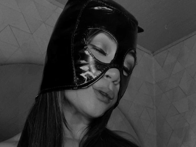 SteffyMiller wearing a shiny black latex mask with cat ears, eyes closed, and a slight smirk, has long dark hair, and is photographed in a black-and-white shot.