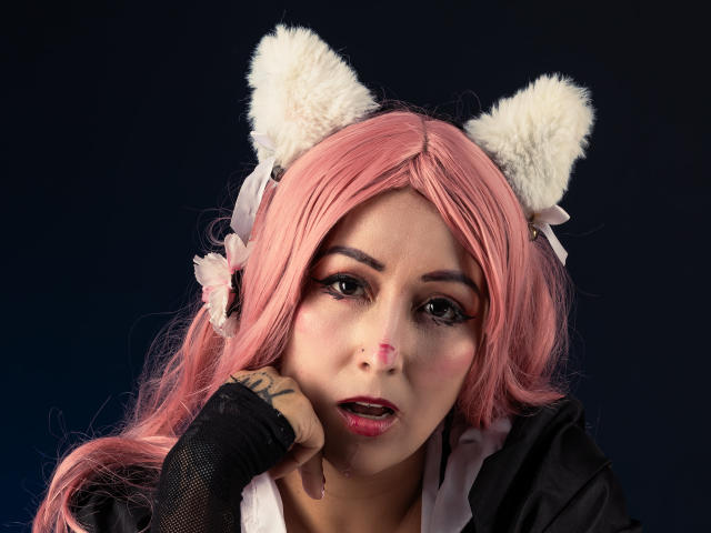 AlanaFerrer with pink hair, white furry cat ears, black fingerless gloves, and a black jacket, has a nose piercing and tattoo on her hand, looking shocked.