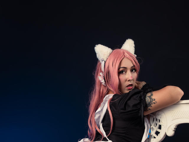 AlanaFerrer with pink hair, white cat ears, black dress, tattoo on right arm, leaning on white chair, looking back with parted lips, dark background. Boobs, cute, sexy.