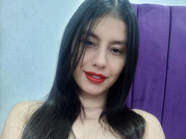 MadissonFontaine with long black hair, red lipstick, and a slight smile, showing her bare shoulders, has a cute face and is looking at the camera.