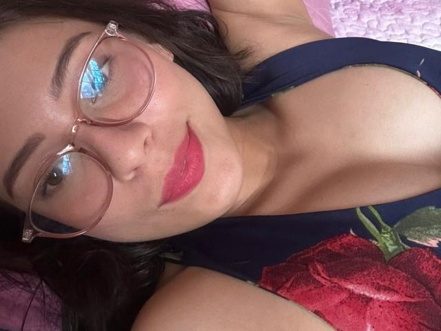 IsabelaPretty with glasses, red lipstick, and a dark blue floral tank top, showing her tits, lying on a pink quilted surface, blue eyes reflecting light.