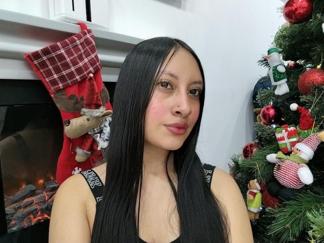 TamyCute with long black hair, pink cheeks, black tank top, standing in front of a Christmas tree and fireplace, smiling slightly. Red plaid stocking with reindeer on mantle.