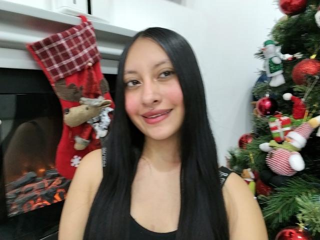 TamyCute has long black hair, light skin, and a small smile, wearing a black top, standing in front of a Christmas tree and fireplace with a red stocking.