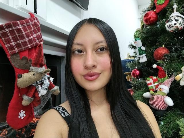 TamyCute with long black hair, pink lips, and black top, stands in front of a decorated Christmas tree, showing a red reindeer stocking. Her tits are covered by the top.