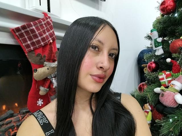 TamyCute has long black hair, brown eyes, pink nose, and a small nose piercing. She's wearing a black tank top with "AVIATOR" text, standing by a decorated Christmas tree.