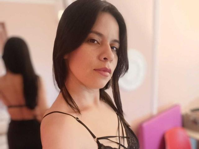 VeronicaHex with black hair, wearing a strappy black top, looking at the camera, her ass in a mirror reflection, soft pink walls, a pink pillow in the background.