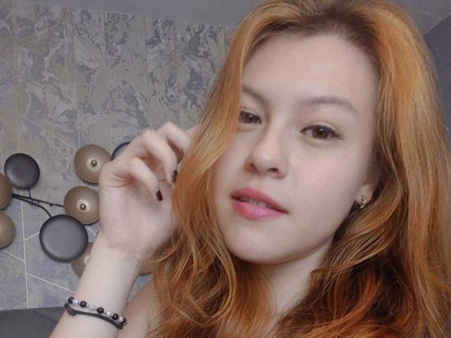 TaylorCarpert has pale skin, red hair, and is looking at the camera with slightly parted pink lips. She's touching her hair with black-painted nails.