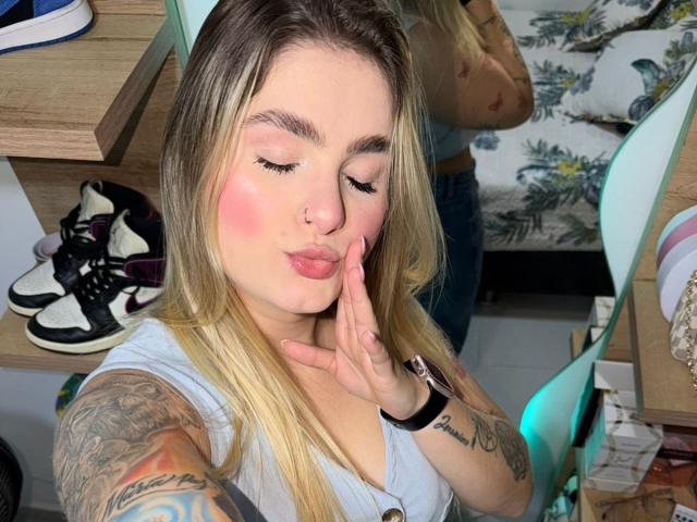 AlannaFoxy with puffy pink cheeks, nose ring, and blonde hair, kissing her hand, showing tattoos on her arm, wearing a gray top, and a black wristwatch.