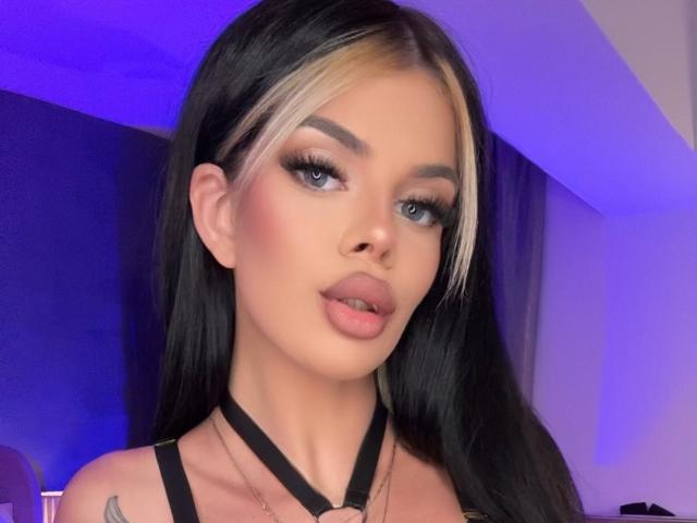 IvyRaven with black and blonde hair, thick eyebrows, long eyelashes, pink lips, black strappy top, small tattoo on left shoulder, blue-purple light, slightly open mouth, looking at camera.