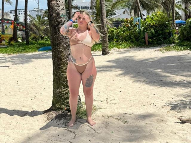 AlannaFoxy stands on a beach, wearing a tan bikini, with tattoos on her arm and thigh. She's drinking from a bottle, her hands near her face.