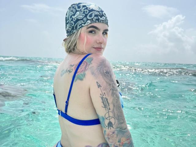 AlannaFoxy in a blue bikini, tattoos on arms and back, bandana on head, standing in clear turquoise water, looking over shoulder, pink blush on cheek, slight smile.