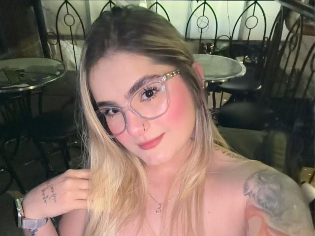 Blond AlannaFoxy with glasses, pink blush, nose ring, tattoos, and a bracelet, smiling at the camera, showing bare shoulders, in a dimly lit café.
