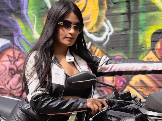 JulietaRizzo with long black hair, wearing aviator sunglasses, a white and black leather jacket, and black top, riding a motorcycle, gripping the handlebars, showing her tits.