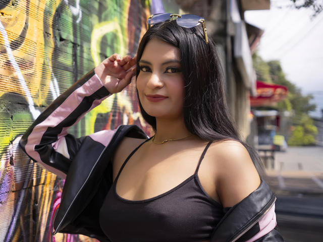 JulietaRizzo with black hair, wearing a black sports bra, and a pink and black track jacket, has her hand near her ear, showing off her small tits.