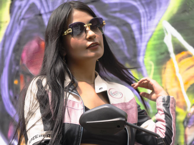 JulietaRizzo with long black hair, wearing sunglasses, a pink and white jacket, and holding a black mirror, looking confident with a slight smile.