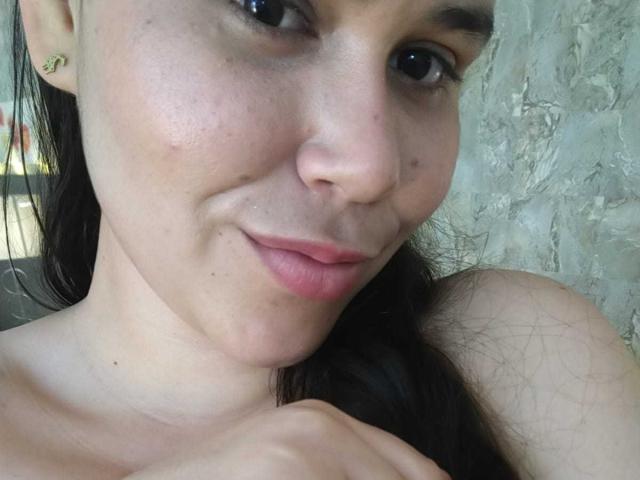 DanTaylor's face, showing light skin with freckles, brown eyes, and pink lips, has a slight smile with a hand near her neck, dark hair, and small gold earrings.