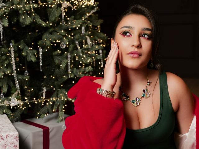 GoldenHear with medium tits, green dress, red lipstick, floral necklace, and bracelet, touching face, Christmas tree with lights in background, red jacket off shoulder, presents on floor.
