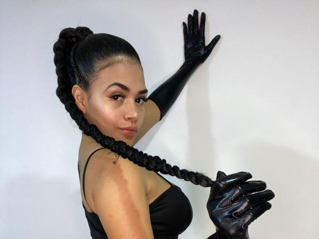 NicolleVilla with long black braids, wearing black gloves, touching wall, showing side-boob, looking over shoulder, glossy lips, dark eyebrows, white background, standing, confident pose.