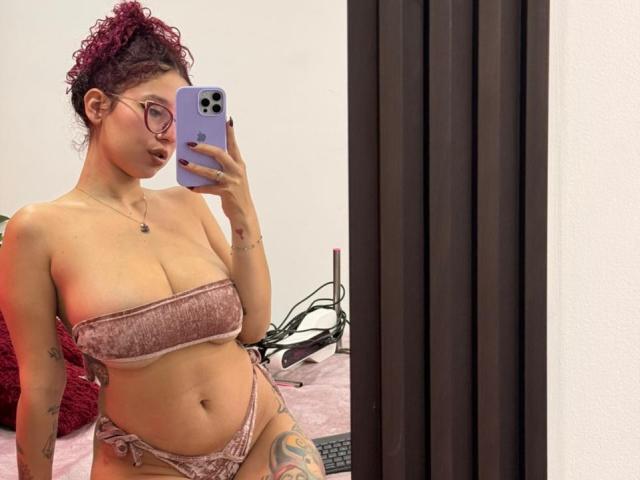 ValeryScot with curly red hair, glasses, and pink velvet bikini, holding a phone, showing her boobs, ass, and a colorful thigh tattoo, taking a mirror selfie.