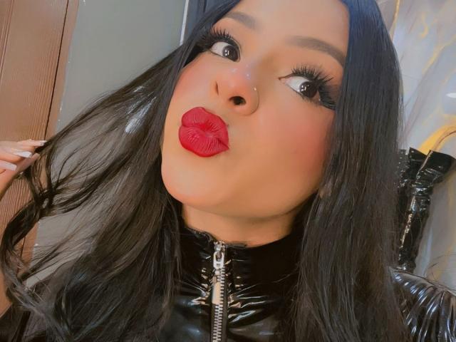 LindaFrancoo with long black hair, red lipstick, and black glossy zip-up jacket, pouting her full lips and looking up with dark, thickly lined eyes and long lashes.