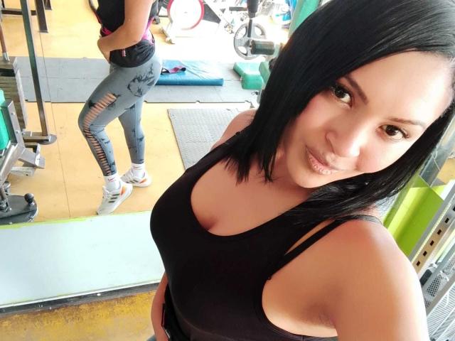EstrellaSmmith, con i capelli neri a metà schiena, indossa un reggiseno sportivo nero, mostrando il seno, e leggings grigi strappati, è in piedi in una palestra e sorride alla fotocamera..