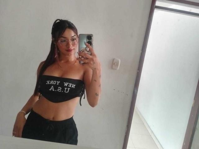 SashaHans with glasses, long dark hair, black tube top showing her tits, black pants, taking a mirror selfie, holding a phone with a white case.