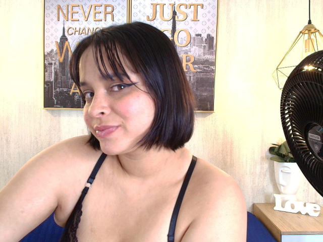 Blueeskyy, con cabello negro corto, lleva un sujetador de encaje negro, tiene senos pequeños, piel de color marrón claro y una ligera sonrisa. A su derecha hay un abanico negro..