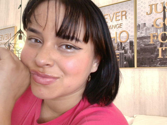 Blueeskyy met een medium bruine huid, zwart steil bob kapsel en roze lippenstift, lachend met glanzende lippen, draagt een roze shirt, op een selfie, met subtiele sproeten en een cat-eye eyeliner..