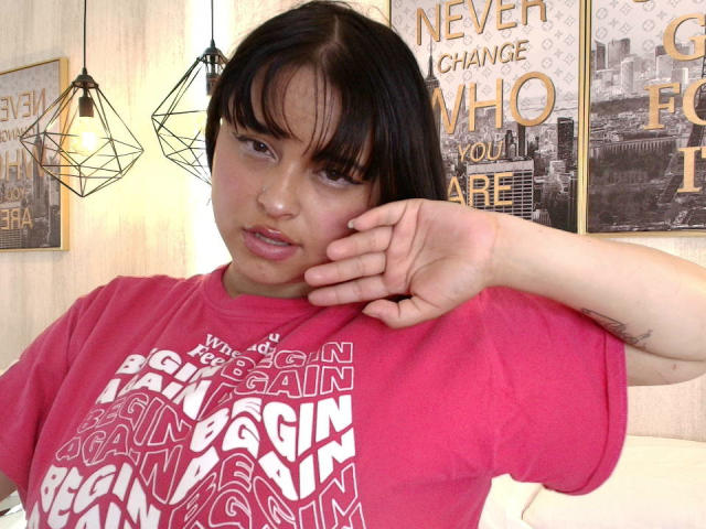 Blueeskyy, met zwart haar en een pony, draagt een rood T-shirt met de tekst "New York City" en heeft haar hand in de buurt van haar gezicht, waarbij een tatoeage op haar rechterarm zichtbaar is..