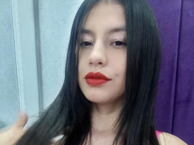 Madisson Fontaine tiene el cabello negro y liso, lápiz labial rojo y una pequeña marca de nacimiento debajo del ojo derecho. Está mirando ligeramente hacia un lado con una expresión neutra..