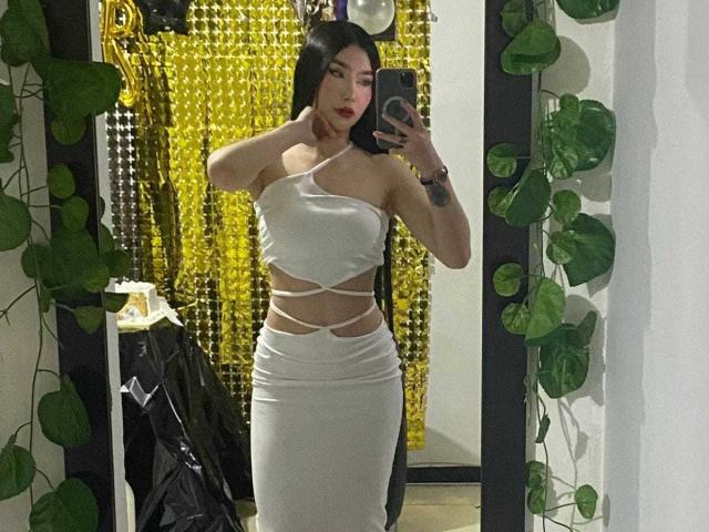 BellaDalthon in a white, one-shoulder, cut-out dress, showing her small tits and slim ass, taking a mirror selfie with green leafy plants around. She has black hair and red lipstick.