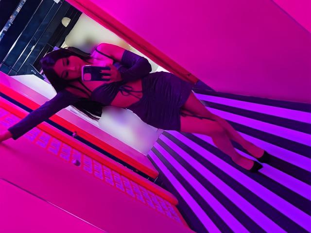 Zawora in a tight purple dress with long sleeves, showing her ass and tits, taking a mirror selfie in a pink and purple lit room, wearing black heels.