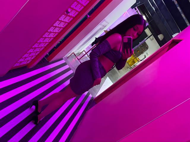 Zawora with long black hair, purple off-shoulder top, and matching purple skirt, holding phone, standing in pink and purple striped room, wearing black high heels.