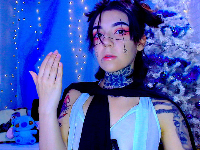 Lannie Gonzales, con cabello negro, gafas, sombra de ojos rosa, blusa blanca, capa negra, tatuajes y bufanda azul, con las manos juntas, al lado de un árbol de Navidad iluminado con luces azules y blancas..
