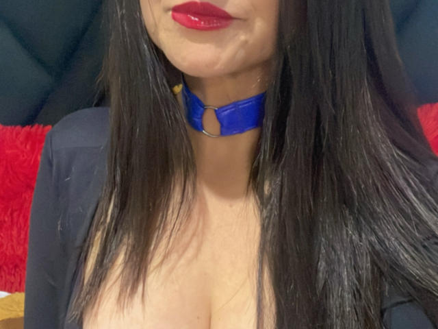 Close-up of KynkyFetish's upper body. She has long, straight black hair, full red lips, and a blue choker with a small ring in the center. Her cleavage is visible, suggesting large breasts. She wears a black garment, partially open. The background includes a red, fuzzy texture and black, cushioned material. The image focuses on her face, lips, and choker. The photo has a casual, slightly blurry quality.