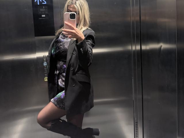 Lellie with blonde hair, wearing a black blazer, floral dress, and black boots, taking a mirror selfie in a metallic elevator, showing her knee up and phone in hand.