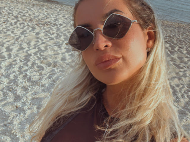 Lellie with blonde hair, wearing dark sunglasses and a black top, has pouty lips and a slightly smug expression on a pebbly beach.