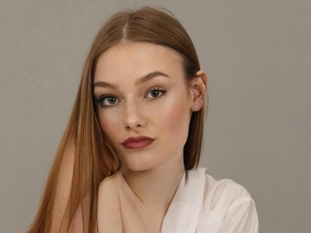 Arieell has long brown hair, a nose ring, and wears a white shirt. Her brown eyes, dark eyeliner, and red lipstick are prominent. She's looking at the camera with a slight smile.