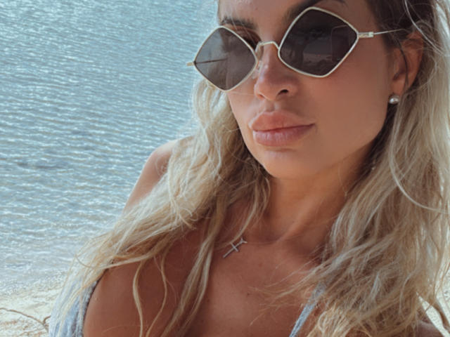 Lellie with long blonde hair, wearing hexagonal sunglasses, a cross necklace, and a grey bikini top, showing her boobs, standing by a water body.