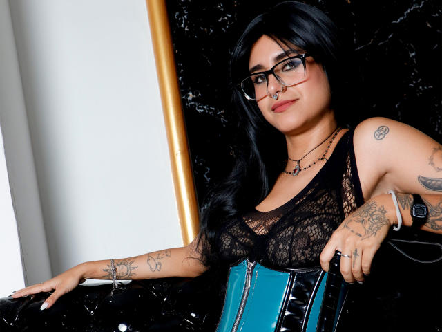 SamanthaGio with black lace top, blue corset, tattoos, glasses, nose ring, and wristwatch, leaning on black chair, showing her tits and ass, with long black hair.