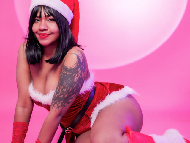 MaddyMagic with black hair, red Santa hat, red lace lingerie, white fur trim, red gloves, tattoo on left arm, smiling, kneeling, showing large tits, ass, pink background.