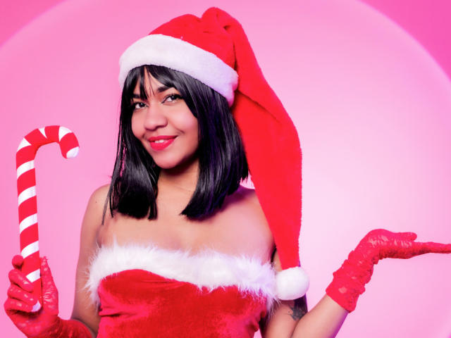 MaddyMagic with black bob, red Santa hat, red velvet top, white fur trim, red gloves, holding candy cane, smiling, pink background. Boobs, cute, sexy, festive, flirty.
